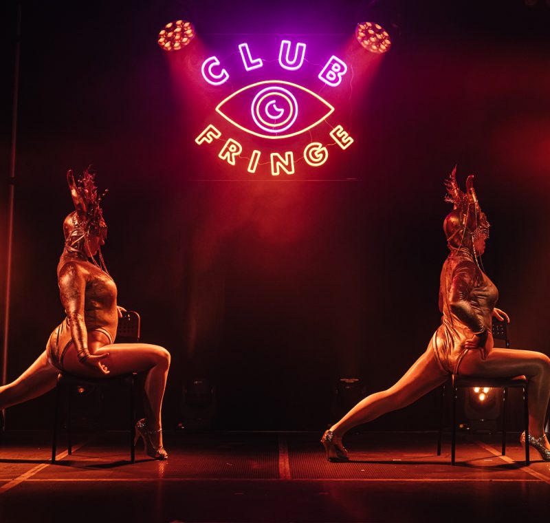 Two burlesque performers on stage in costumes and large head-dresses, sitting sideways on chairs