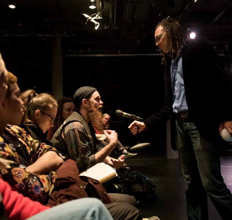 A man wearing glasses and dreadlocks holds a microphone towards a man with a beard, who is speaking into it, surrounded by other people in a theatre space