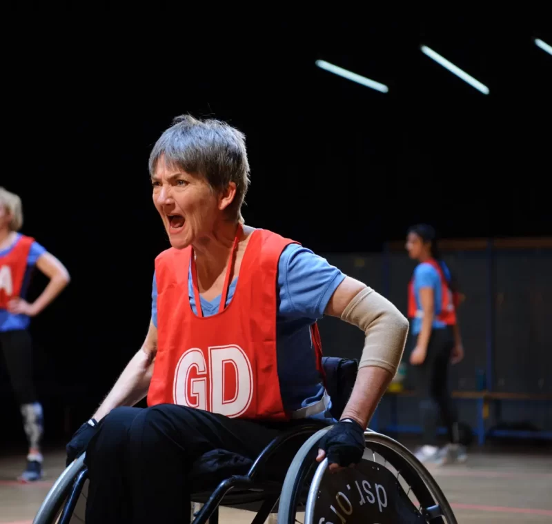 A woman sitting in a wheelchair, wearing a netball bib, and calling out