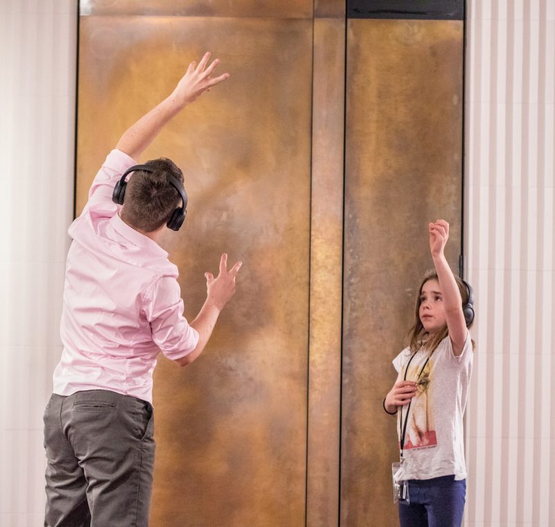 A man and child, both wearing headphones, face each other and reach one hand to the sky as if mirroring each other.