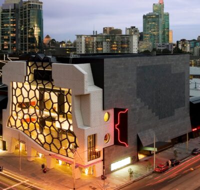 Elevated street view of a large building on a Melbourne street corner at dusk. One side of the building is dark grey, the other side is light beige with honeycomb shaped windows set into the middle of the wall. The building is lit from the inside.