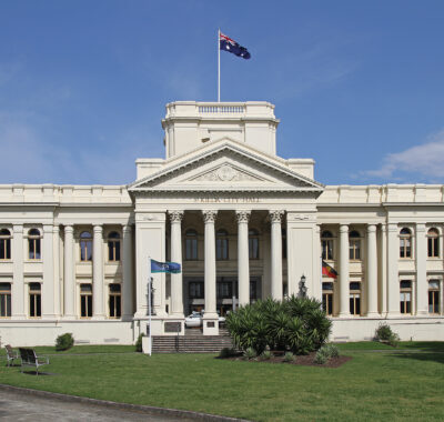 Neoclassical style building with light beige exterior. The sky is blue and ther eis green grass and some small shrubs in front of the building