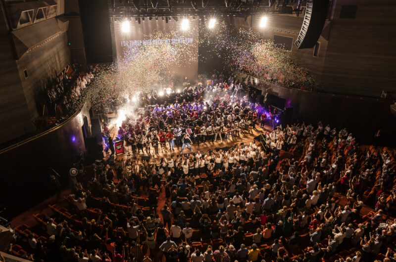 Birds eye view of a theatre full of people giving a standing ovation. On stage there are a large number of people standing in rows playing guitars. Confetti is falling from the ceiling.