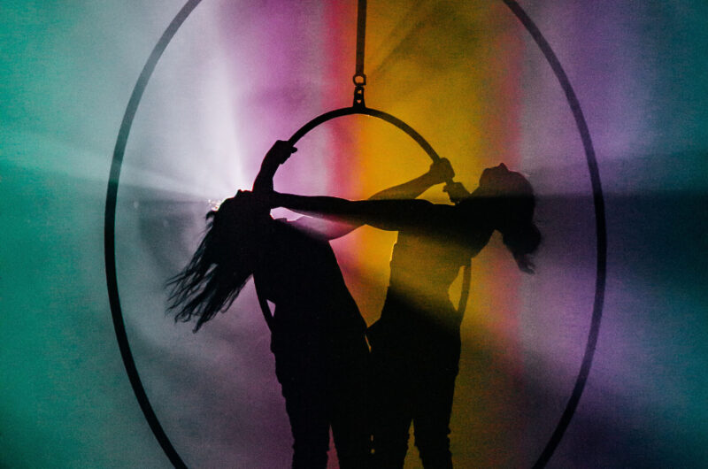 Two women stand in a large hoop suspended from the ceiling. Theu are silhouetted against rainbow coloured lights projected around them