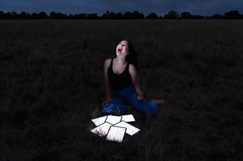 A woman sits in a field looking up at the sky and screaming. The sky is dark as if it is night time. There are 6 small white rectangles of light arranged in an pile in front of the woman shining some light onto her face.