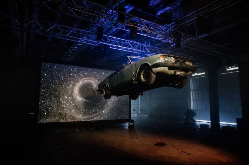 AN old fashioned convertible car is suspended in the air inside a warehouse. Behind the car an image of stars in outer space is projected onto a large screen
