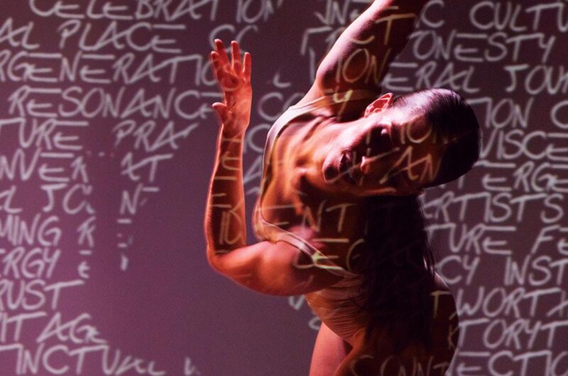A dancer twists sideways and reaches up in front of a mauve background. There is writing projected onto the background and some parts of the dancer's body.