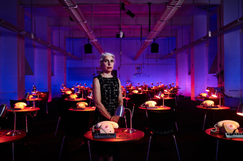 A woman stands in the centre of a purple dimly lit room. She is wearing a black short sleeve dress and looking directly at the camera with a serious expression. She is surrounded by rows of small round tables. On each table there is a lamp and a rotary telephone.