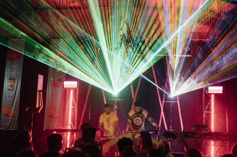 Two men stand on a stage in a warehouse surrounded bvy bright party lights and music equipment. In the foreground a crowd of people are dancing.
