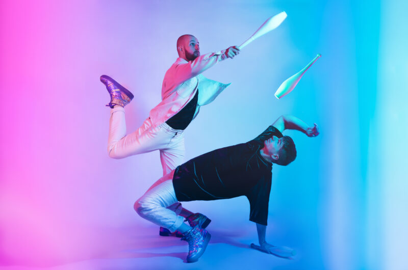 Two men stand in an empty space lit with purple and blue lights. One man is bent over backwards with one hand on the gorund. The other man is reaching forward over him, balanced on one leg. The men are each throwing one juggling club through the air
