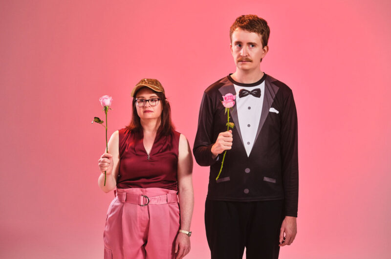 A man and woman stand side by side in front of a pink background. The man is wearing black trousers and a long sleeve top printed to look like a tuxedo. The woman is wearing pink trousers, a red short sleeve top and a sparkly gold baseball cap. Both people are holding single pink rose in their right hand.