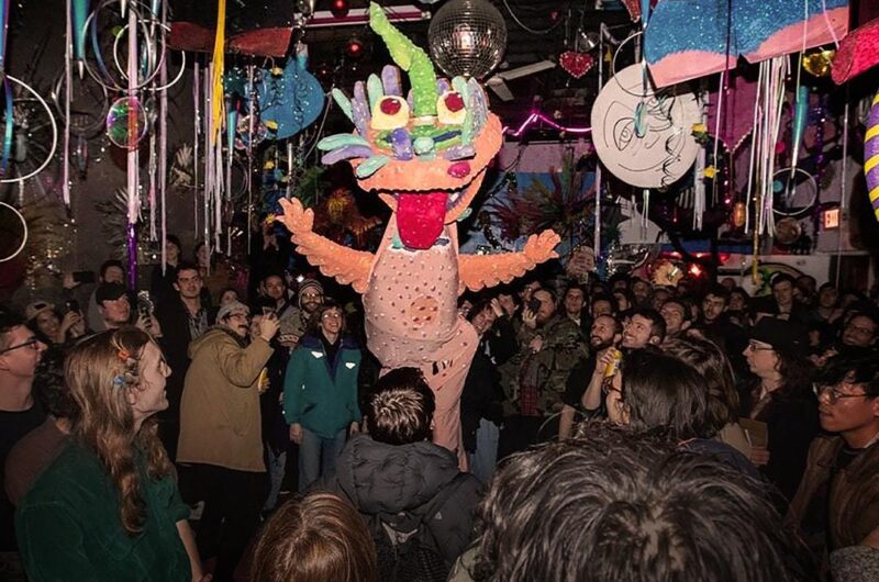 A large number of people are gathered at a party with colourful decorations hanging from the ceiling. In the centre of the people is someone wearing a giant dragon costume. The costume is coloured pink, purple, green and blue with a red tongue poking out of the dragons mouth.