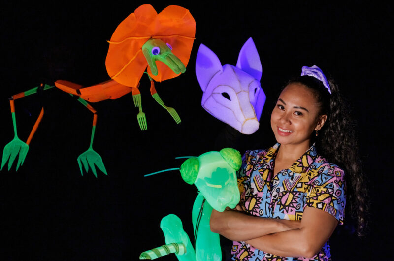 A woman stands in front of a black background, smiling at the camera. She is wearing a colourful patterned shirt and her hair is in a ponytail. She is surrounded by three illuminated puppets in the shapes of animals.